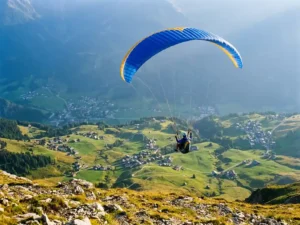 Paraglider zweeft boven een Alpendal met kleurrijke vleugel tegen kobaltblauwe lucht, groene weiden en dorpjes diep beneden.