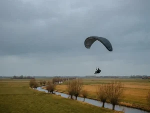 Paraglider vliegt laag over een Hollands polderlandschap in de herfst, met gouden velden en kale wilgen langs een kanaal.