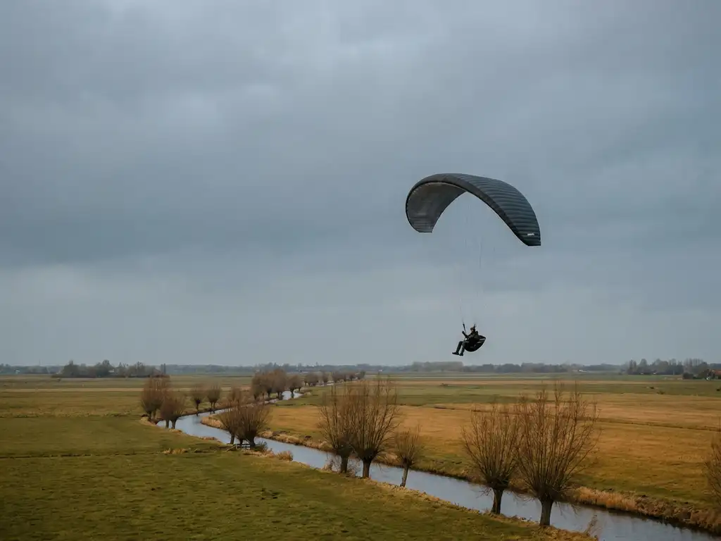 Paraglider vliegt laag over een Hollands polderlandschap in de herfst, met gouden velden en kale wilgen langs een kanaal.