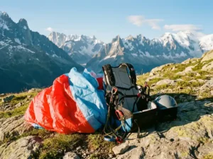Paragliding starterspakket met harnas, helm en gevouwen kleurrijke vleugel op een rotsachtig Alpenweiland, bergtoppen op de achtergrond.