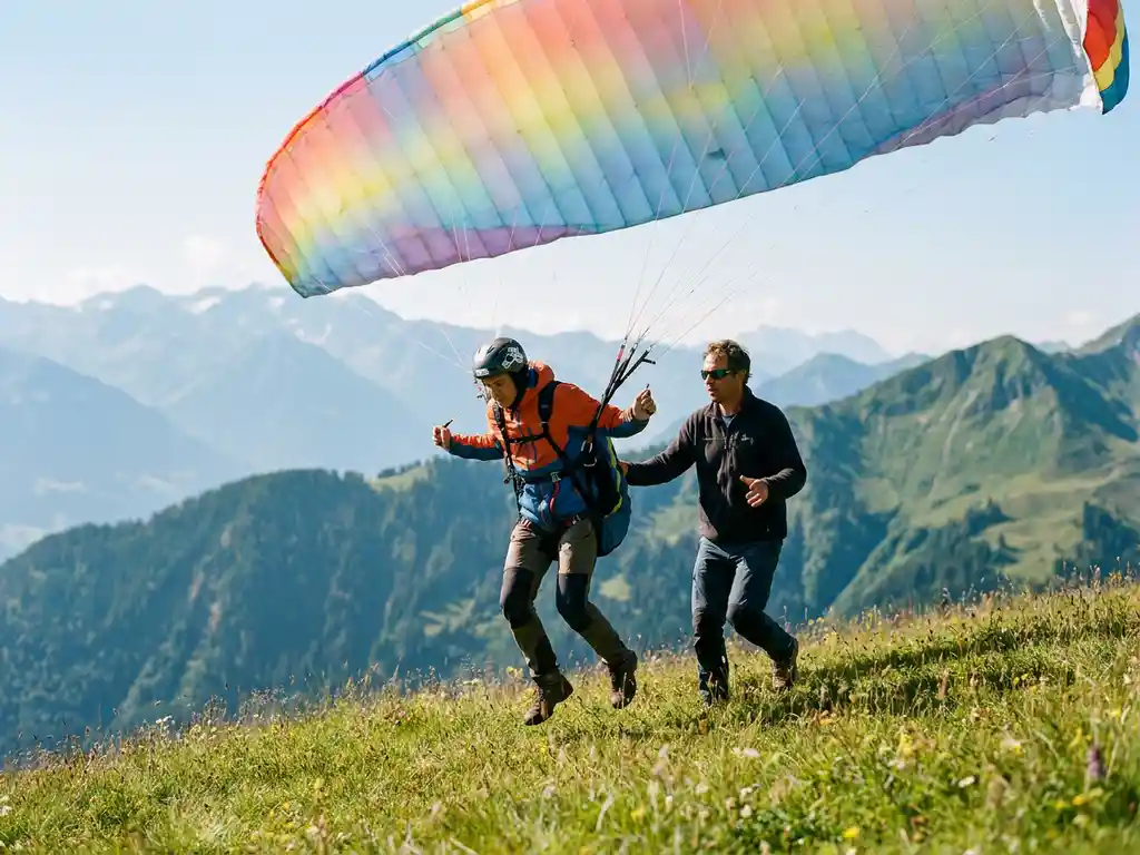 Student paraglider lanceert van een grazige Alpenhelling, kleurrijke vleugel boven hoofd, instructeur begeleidt de eerste vlucht met bergpanorama op de achtergrond.
